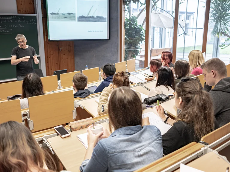 Vorlesungssaal, viele Studierende, Dozentin vor der Tafel