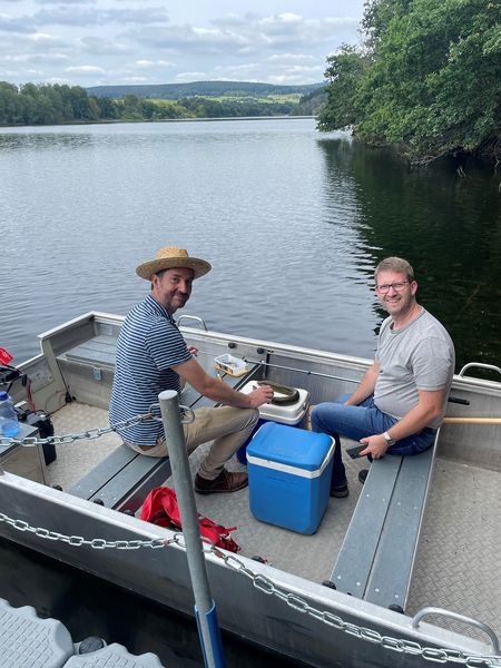 Prof. Dr. Stoll & Prof. Dr. Didas sitzen in einem Boot. Vor Ihnen liegt ein Aal auf einem Tisch.