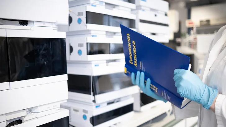 Titelbild: Labor, Person in Laborkittel mit Buch vor Laborgeräten